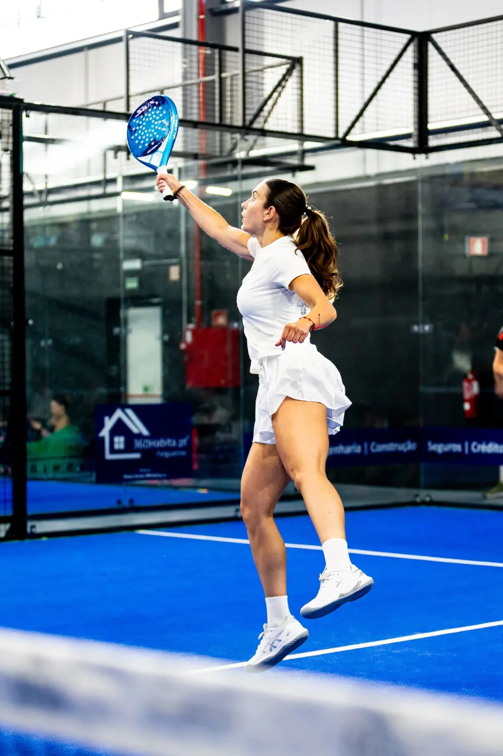 Jogadora a esticar-se para um smash num campo azul do Hype Padel Club.