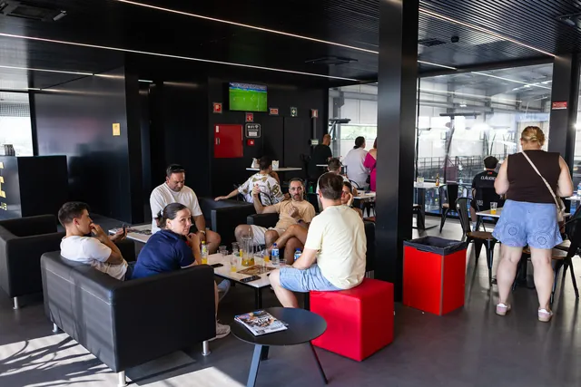 Sofás pretos no bar, com gente à conversa e os campos por trás do vidro.